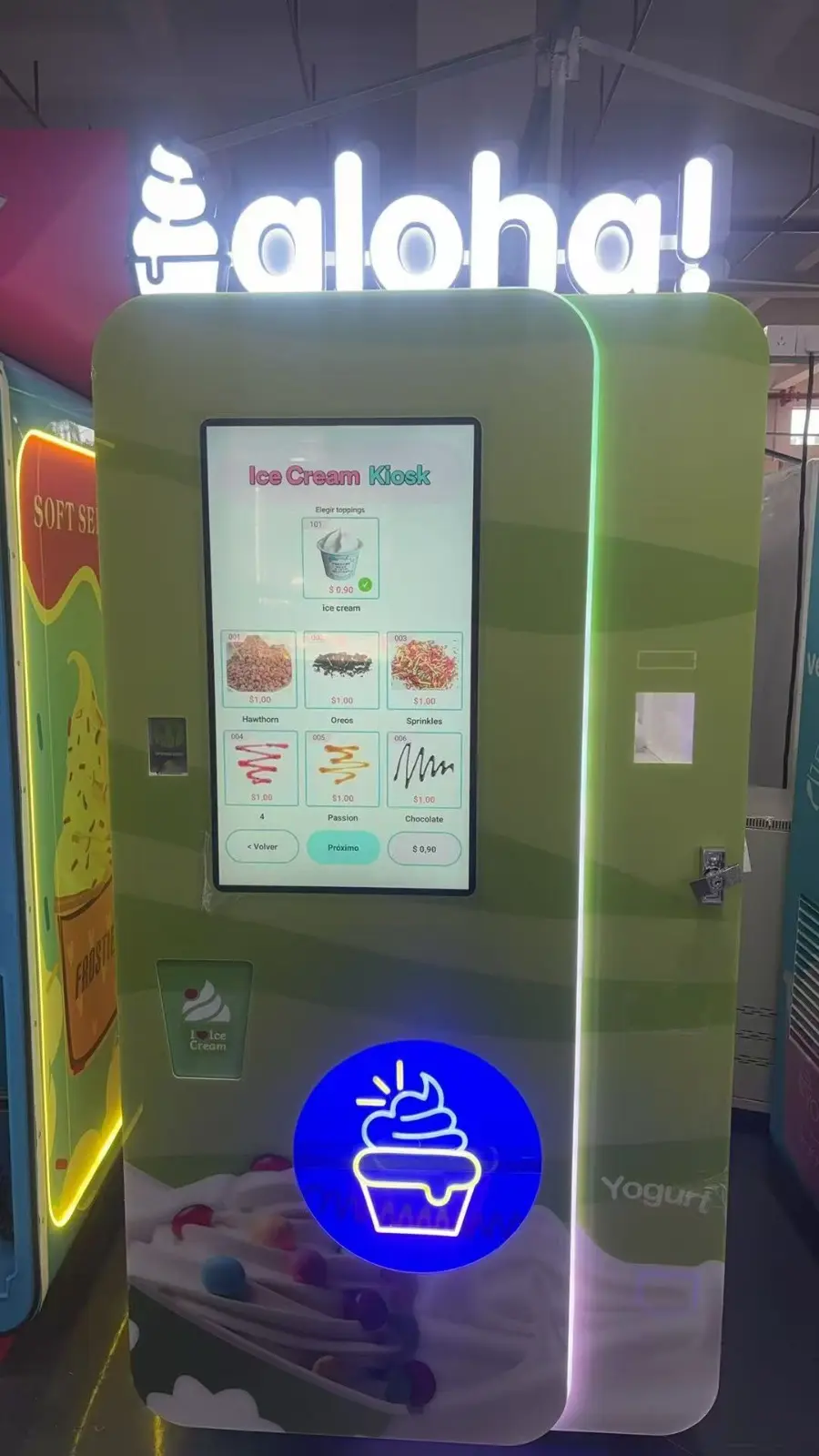 Ein Automat desselben Modells, ganzes Gerät: grünes Gehäuse mit hochformatigem Touchscreen, darüber die Leuchtschrift des Kunden, davor die Becherausgabe.